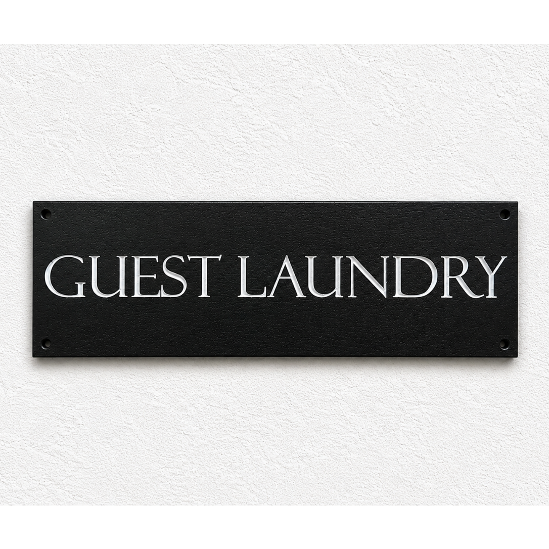 A rectangular black sign with the words 'GUEST LAUNDRY' in white serif lettering, mounted on a textured white wall.