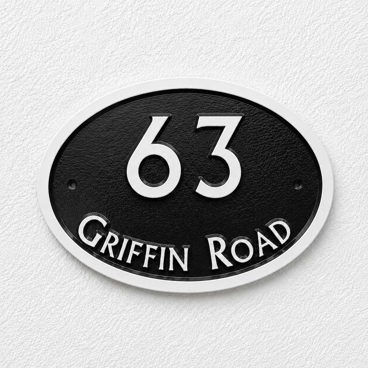 An oval black house number plaque with a white border, featuring the number 63 in white and the text 'GRIFFIN ROAD' in white along the bottom, mounted on a white textured wall.