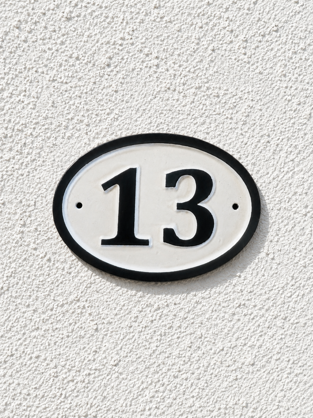 An oval house number plaque with the number 13 in black on a white background, mounted on a textured white stucco wall.