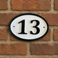An oval white house number plaque with a black border and black numerals reading '13', mounted on a red-brown brick wall.