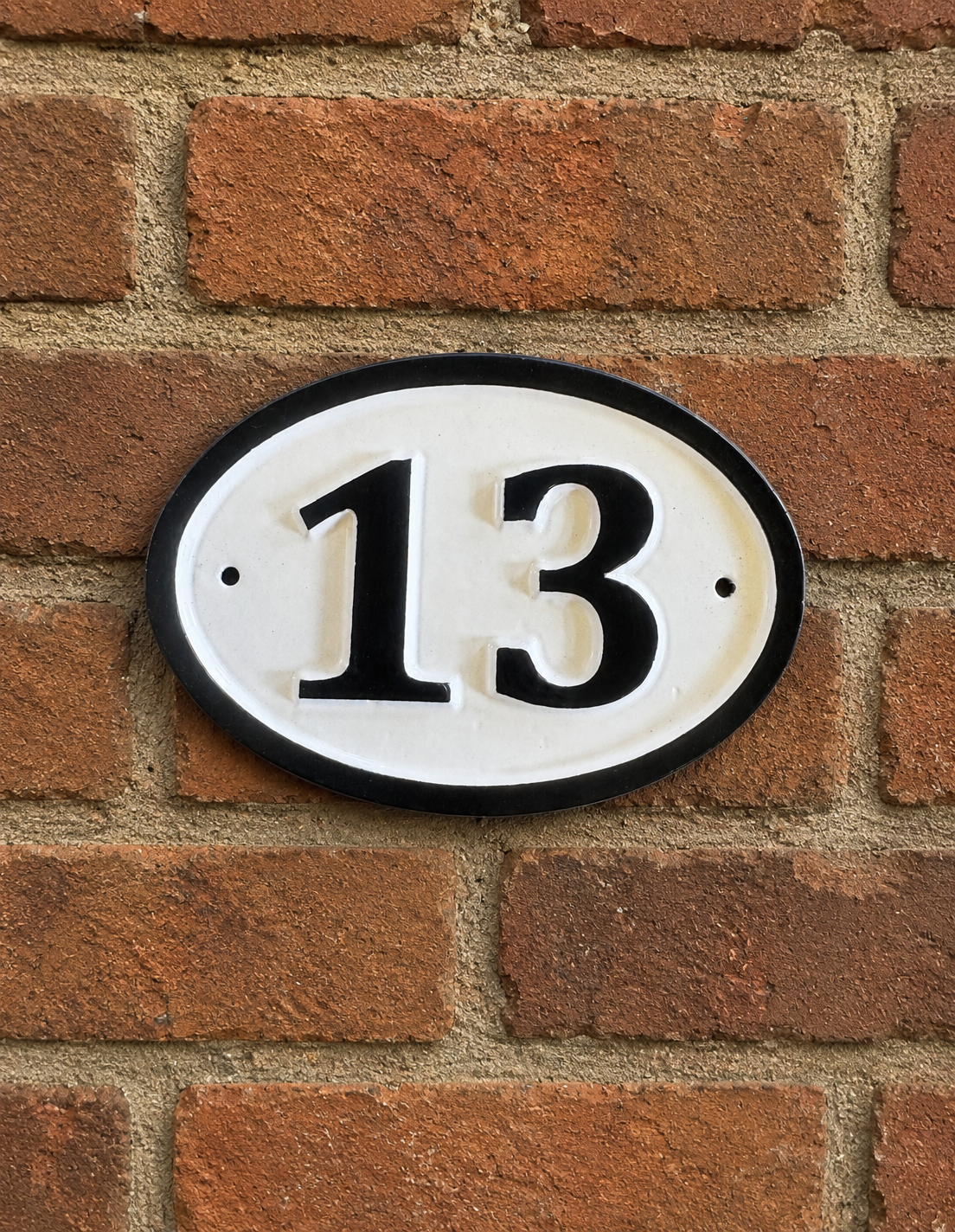 An oval white house number plaque with a black border and black numerals reading '13', mounted on a red-brown brick wall.