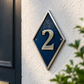 A diamond-shaped house number 2 wall plaque with a textured dark blue center and a silver metallic border, mounted on a white textured stucco wall next to a dark door with green foliage in the foreground.