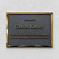 A rectangular memorial plaque with a textured dark gray center and polished gold-toned border, mounted on a white textured wall. The plaque reads 'In memory of Gaston Khoury' with additional engraved text below.