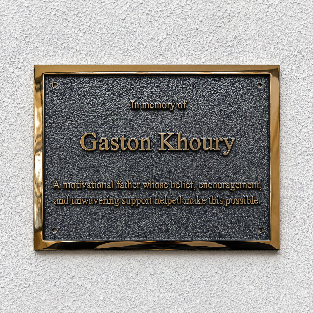A rectangular memorial plaque with a textured dark gray center and polished gold-toned border, mounted on a white textured wall. The plaque reads 'In memory of Gaston Khoury' with additional engraved text below.
