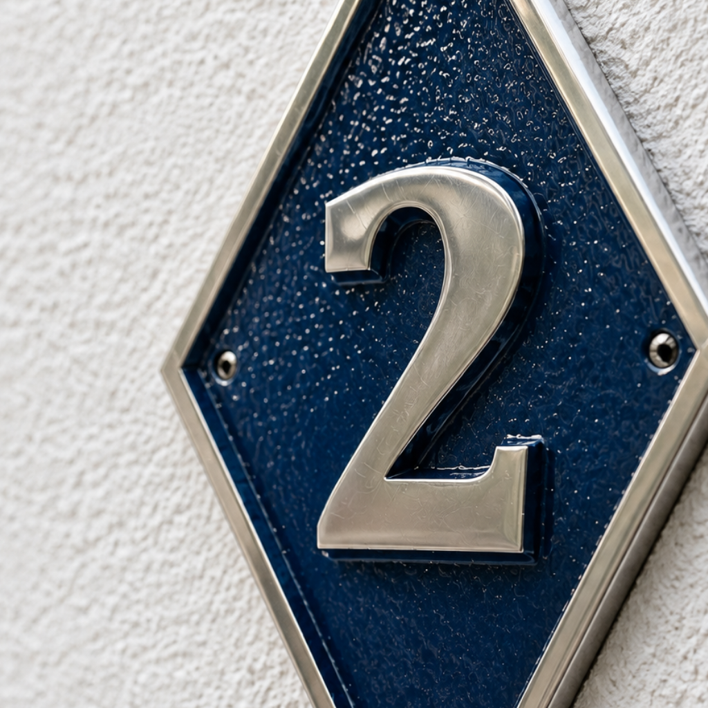 A close-up of a house number '2' plaque on a textured white wall. The plaque features a dark blue textured center, a silver metallic border, and a raised silver number '2'.