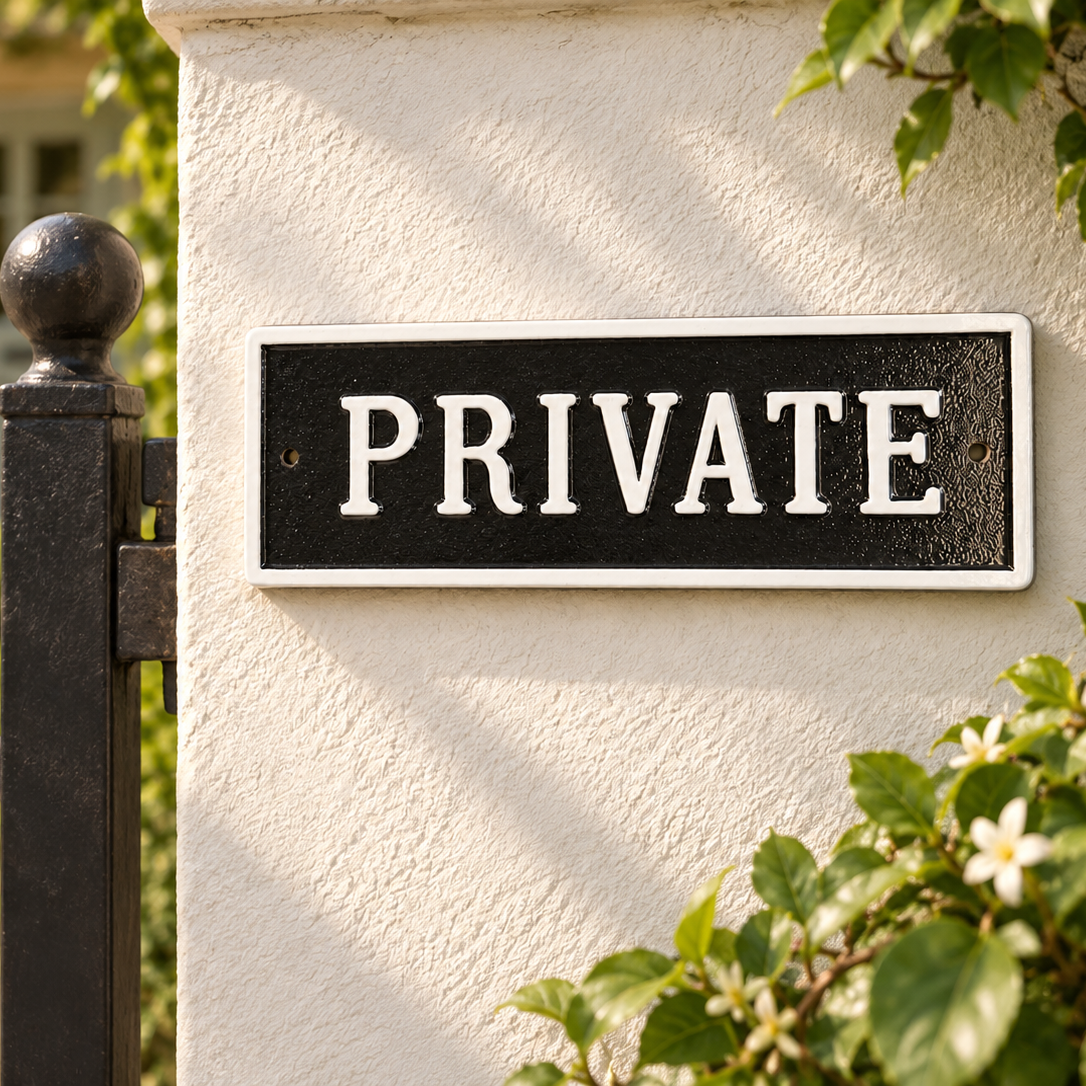 A rectangular black sign with white serif text reading “PRIVATE,” mounted on a cream stucco wall beside a black metal gate, with green foliage and a blurred house entrance in the background.