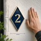 A diamond-shaped house number plaque with a textured dark blue face and silver number '2', mounted on a white stucco wall. A person's hand is touching the plaque, and dimension lines show 184mm height and 110mm width.