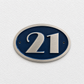 An oval house number plaque featuring the number 21 in brushed silver metal over a deep blue background, mounted on a textured white wall.