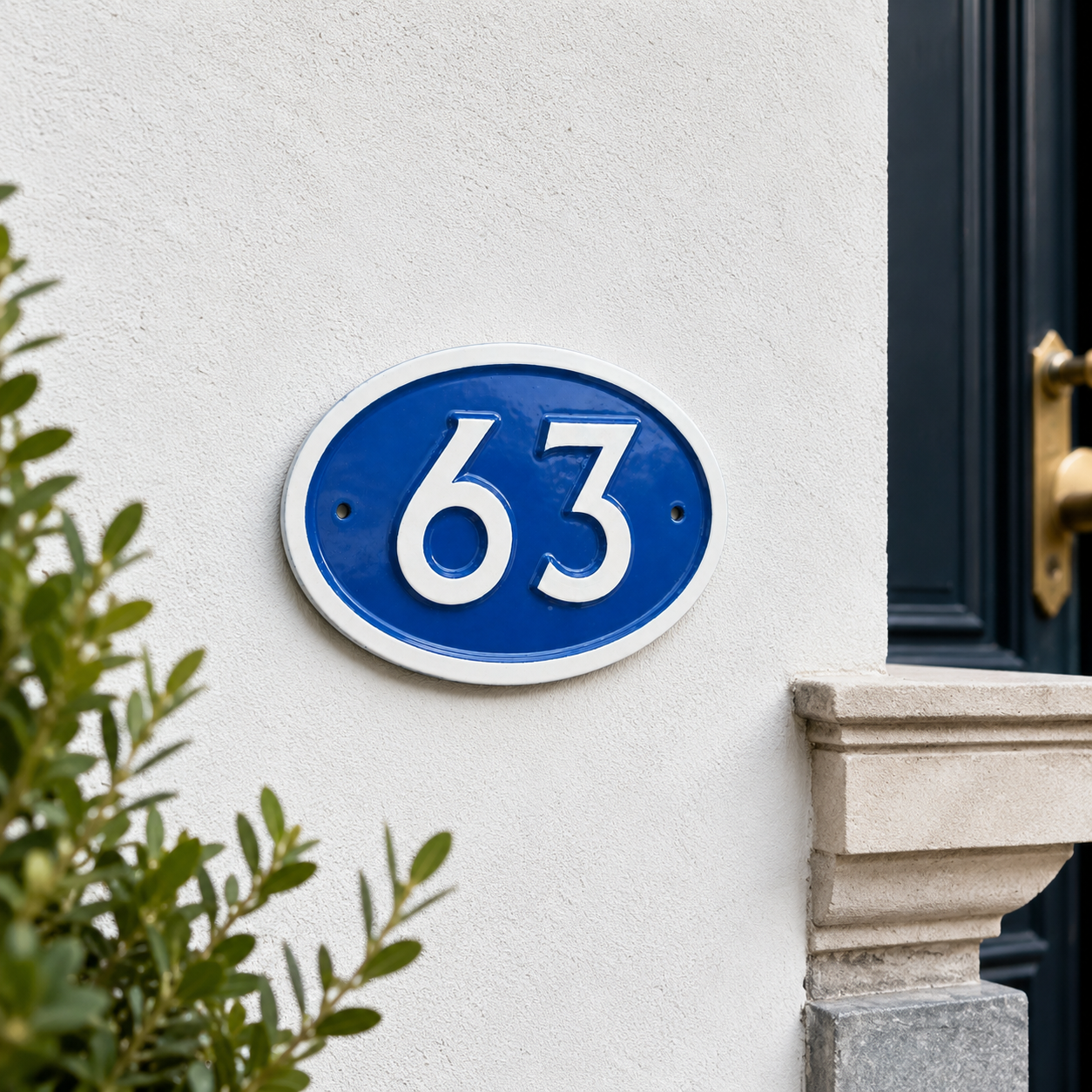 A blue oval house number plaque with white lettering reading '63' mounted on a white stucco wall next to a dark door and green foliage.