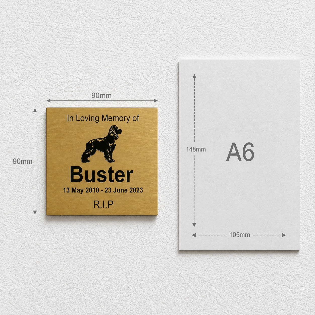 A square engraved pet memorial plaque shown next to an A6 card size reference on a textured white wall. The plaque is a warm metallic gold color with black engraved text and a black dog silhouette.