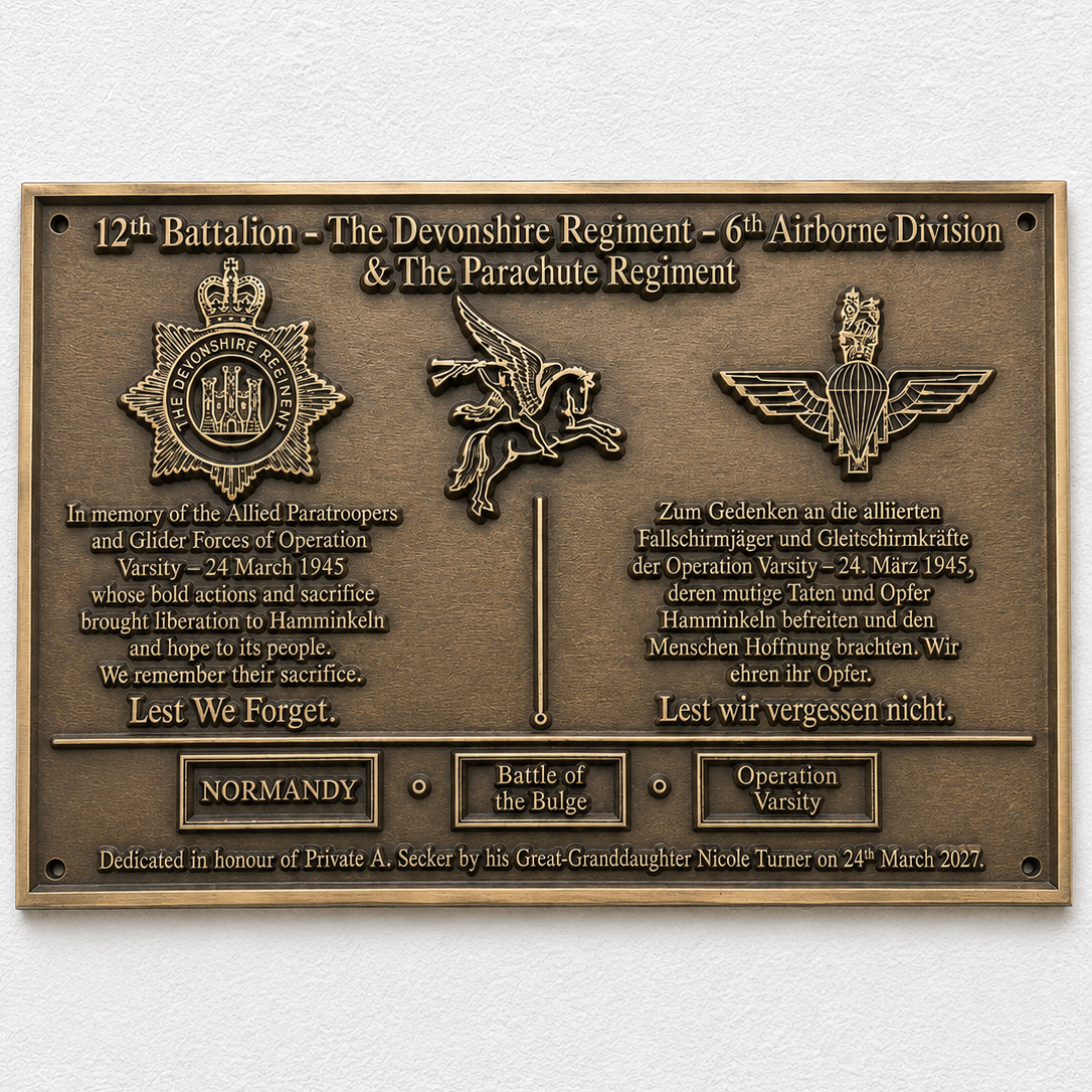A rectangular bronze military commemorative plaque mounted on a white textured wall. The plaque features embossed military insignia, including a crown, a winged figure, and a parachute emblem, along with bilingual text honoring the 12th Battalion, The Devonshire Regiment, and the 6th Airborne Division.