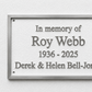 A rectangular memorial plaque with a silver metal frame and raised gray lettering on a textured white background, mounted on a light wall. The text reads: 'In memory of Roy Webb 1936 - 2025 Derek & Helen Bell-Jones'.