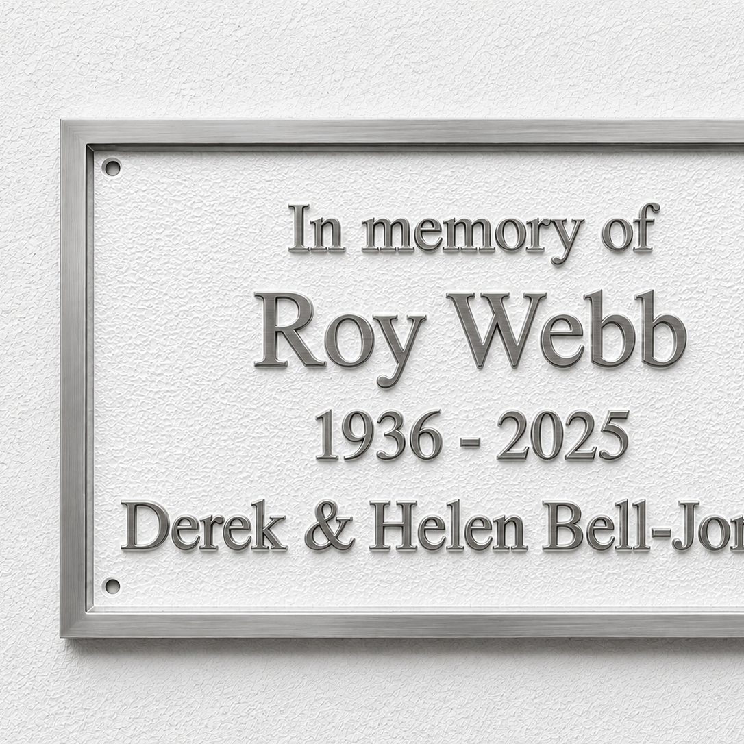 A rectangular memorial plaque with a silver metal frame and raised gray lettering on a textured white background, mounted on a light wall. The text reads: 'In memory of Roy Webb 1936 - 2025 Derek & Helen Bell-Jones'.