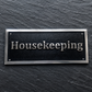A rectangular black plaque with silver lettering reading 'Housekeeping' and a silver border, mounted on a dark slate-textured background.