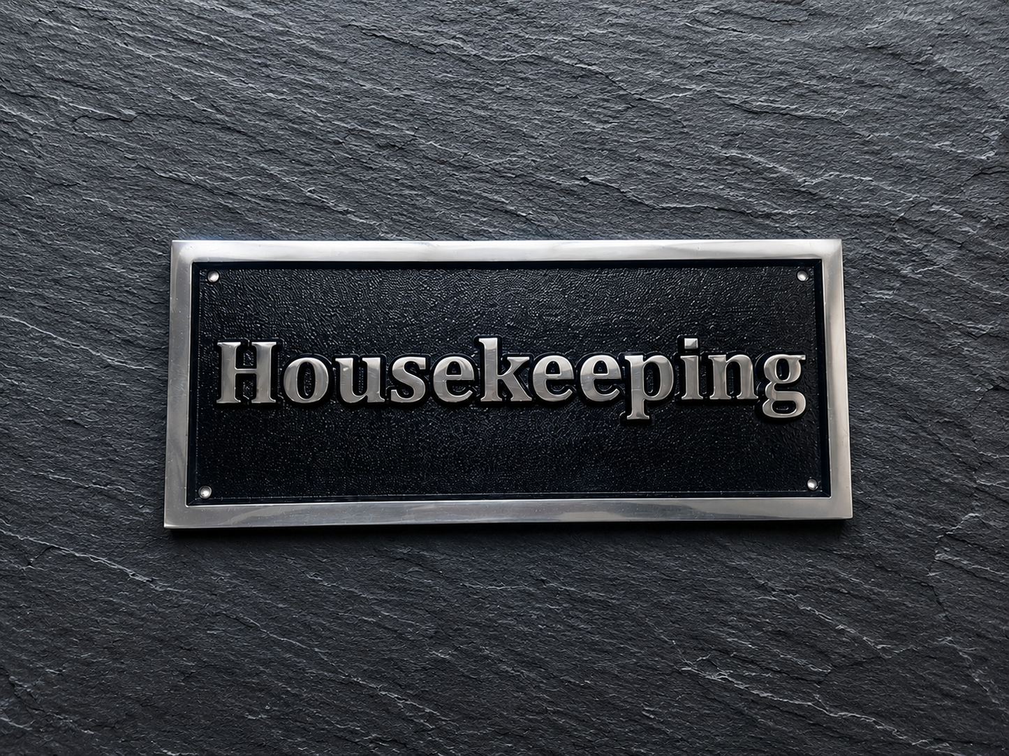 A rectangular black plaque with silver lettering reading 'Housekeeping' and a silver border, mounted on a dark slate-textured background.