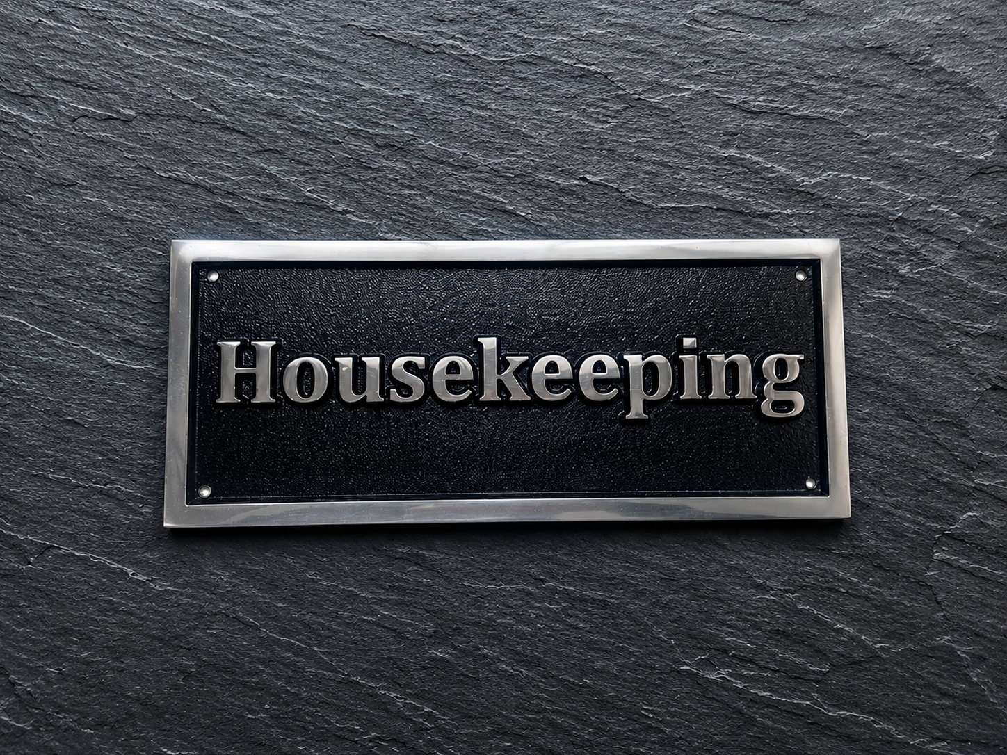 A rectangular black plaque with silver lettering reading 'Housekeeping' and a silver border, mounted on a dark slate-textured background.