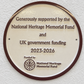 A round commemorative plaque mounted on a textured white wall. The plaque is cream-colored with a dark brown border and raised brown text reading: 'Generously supported by the National Heritage Memorial Fund and UK government funding 2023-2026' with the National Heritage Memorial Fund logo at the bottom.