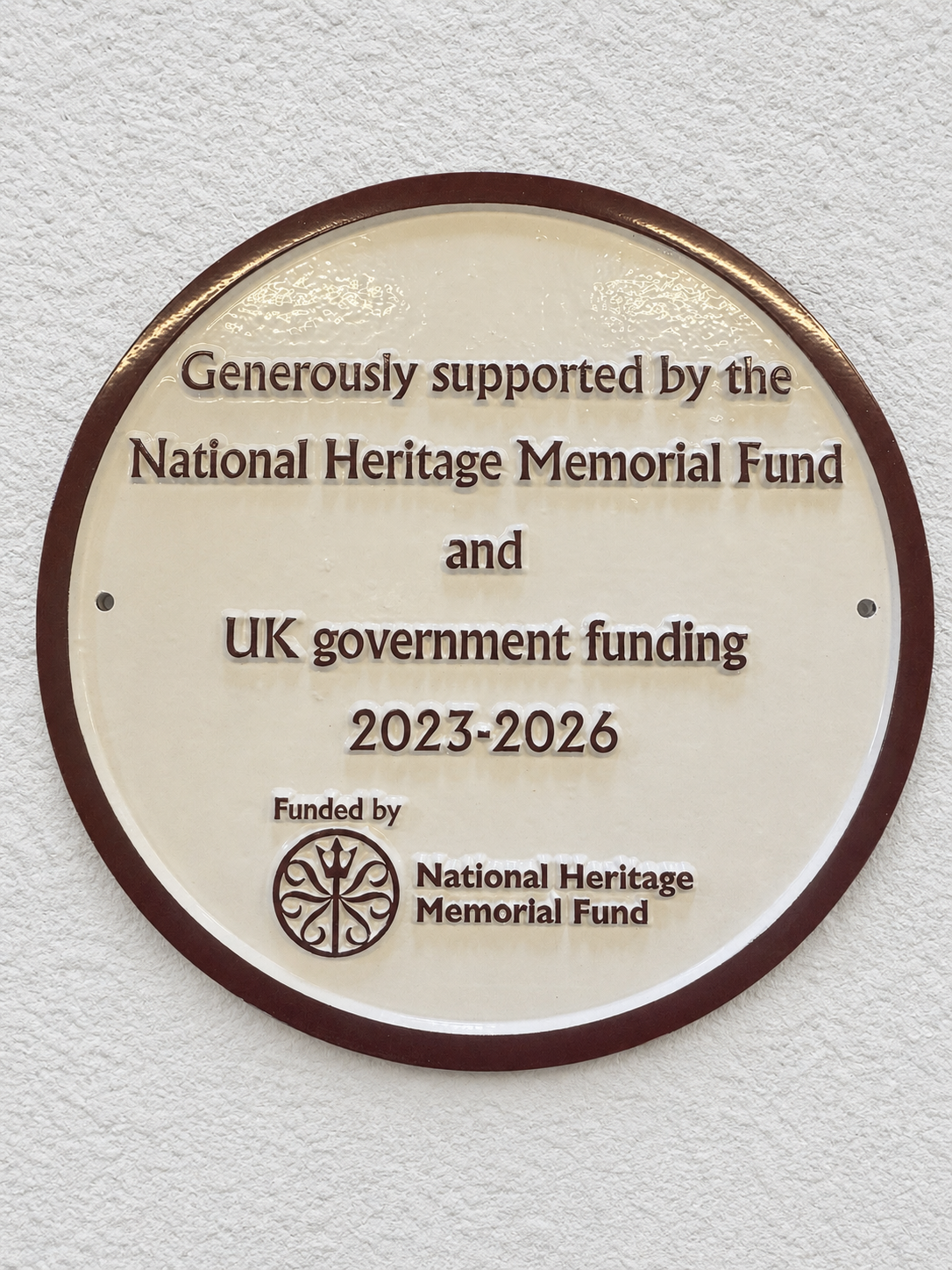 A round commemorative plaque mounted on a textured white wall. The plaque is cream-colored with a dark brown border and raised brown text reading: 'Generously supported by the National Heritage Memorial Fund and UK government funding 2023-2026' with the National Heritage Memorial Fund logo at the bottom.