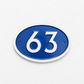 An oval blue and white house number plaque displaying '63' in raised white numerals, mounted on a textured white wall.