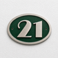 An oval house number plaque featuring the number 21 in brushed silver metal over a textured dark green background, mounted on a white textured wall.