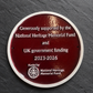 A round dark red commemorative plaque with a white border mounted on a dark slate surface. The plaque features white text acknowledging support from the National Heritage Memorial Fund and UK government funding for the years 2023-2026, along with the National Heritage Memorial Fund logo.