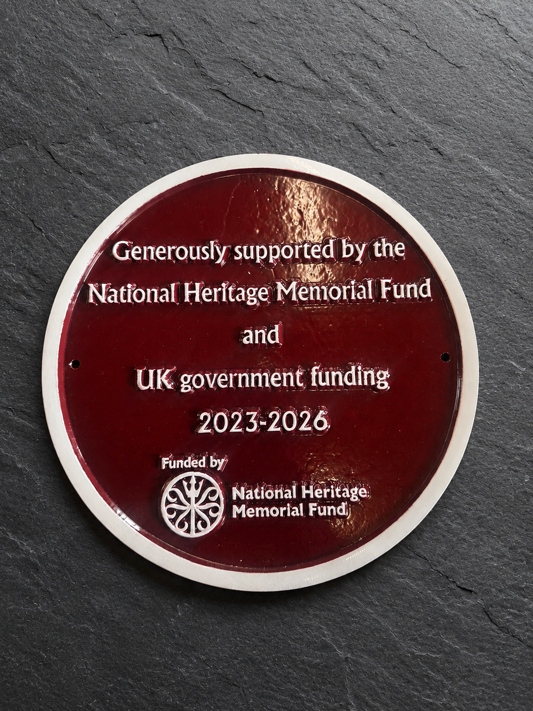 A round dark red commemorative plaque with a white border mounted on a dark slate surface. The plaque features white text acknowledging support from the National Heritage Memorial Fund and UK government funding for the years 2023-2026, along with the National Heritage Memorial Fund logo.