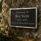 A rectangular memorial plaque mounted on a mossy tree trunk, with flowers at the base. The plaque reads: 'In memory of Roy Webb 1936 - 2025 Derek & Helen Bell-Jones'.