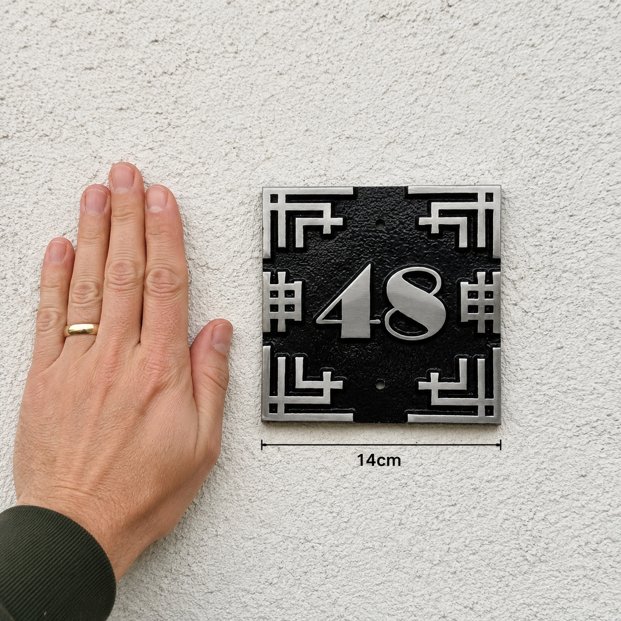 A black and silver house number plaque reading '48' mounted on a textured white wall, shown next to a person's hand for scale with a 14cm dimension marker below.