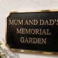 A black rectangular memorial garden sign with gold lettering and a gold border, mounted on a white textured wall. The sign reads 'MUM AND DAD'S MEMORIAL GARDEN'.