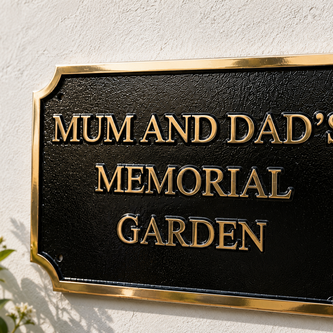 A black rectangular memorial garden sign with gold lettering and a gold border, mounted on a white textured wall. The sign reads 'MUM AND DAD'S MEMORIAL GARDEN'.