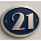 An oval house number plaque featuring the number 21 in brushed silver metal over a dark blue background, mounted on a textured off-white stucco wall.