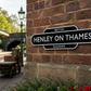 A black and white British Henley on Thames Railways sign mounted on a red-brown brick wall, with a vintage train cart and station platform visible in the background.