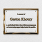 A rectangular gold-framed memorial plaque mounted on a textured white wall. The plaque has a white center with raised gold text reading 'In memory of Gaston Khoury' and a shorter memorial description below.
