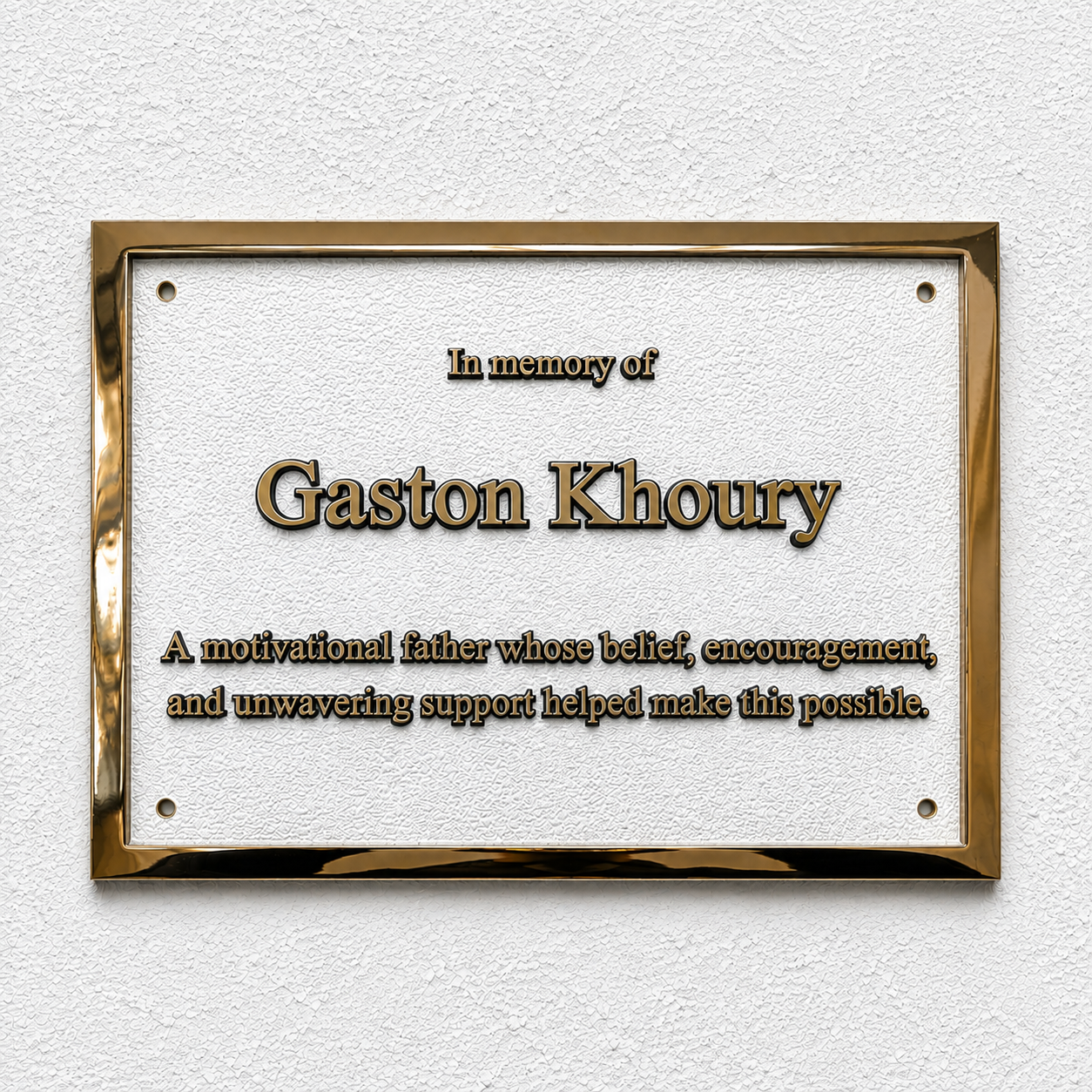 A rectangular gold-framed memorial plaque mounted on a textured white wall. The plaque has a white center with raised gold text reading 'In memory of Gaston Khoury' and a shorter memorial description below.