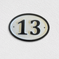 An oval house number plaque displaying '13' in black on a white background with a black border, mounted on a textured white wall.