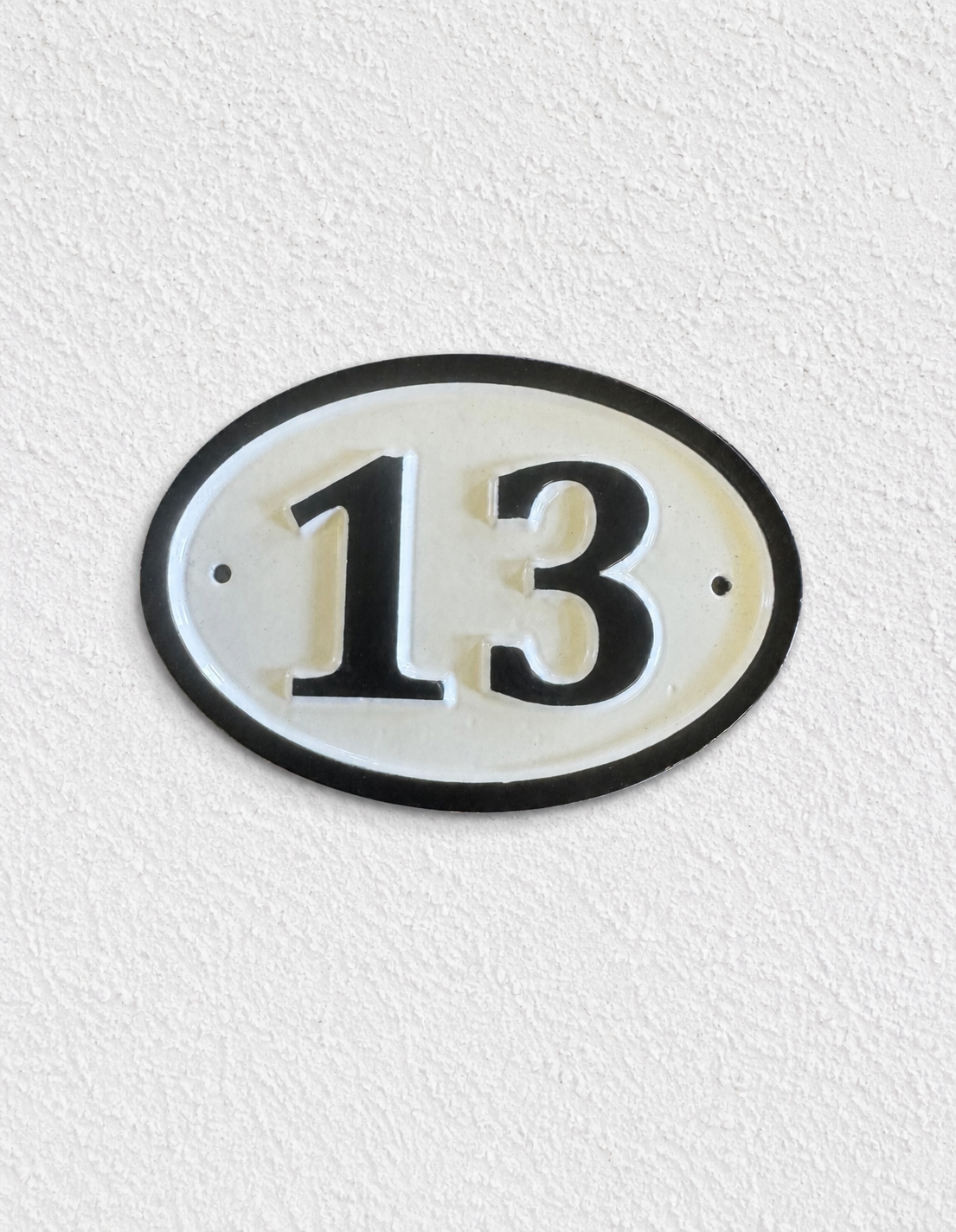 An oval house number plaque displaying '13' in black on a white background with a black border, mounted on a textured white wall.
