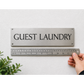 A rectangular brushed-metal guest laundry sign with black lettering is mounted on a white textured wall. A hand holds a metal ruler below the sign to show its size, with a small green plant visible in the lower left corner.
