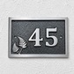 A rectangular house number plaque with the number 45 in raised silver lettering on a dark textured background, bordered by a brushed silver frame. A pair of silver fern leaves is embossed on the left side.