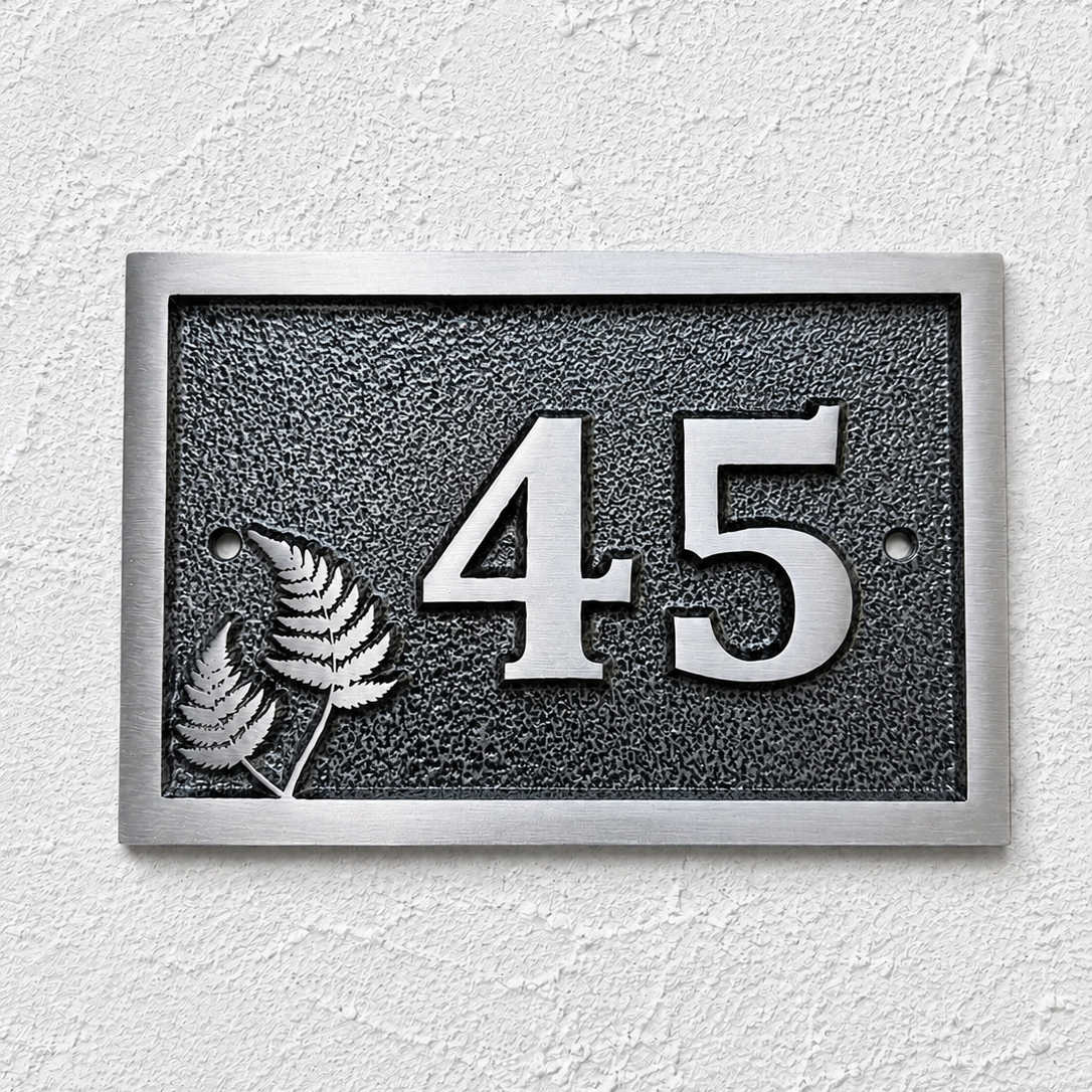 A rectangular house number plaque with the number 45 in raised silver lettering on a dark textured background, bordered by a brushed silver frame. A pair of silver fern leaves is embossed on the left side.