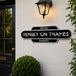 A black and white British Railways station sign reading 'HENLEY ON THAMES' mounted on a cream-colored stucco wall next to a black door and an illuminated wall lamp.