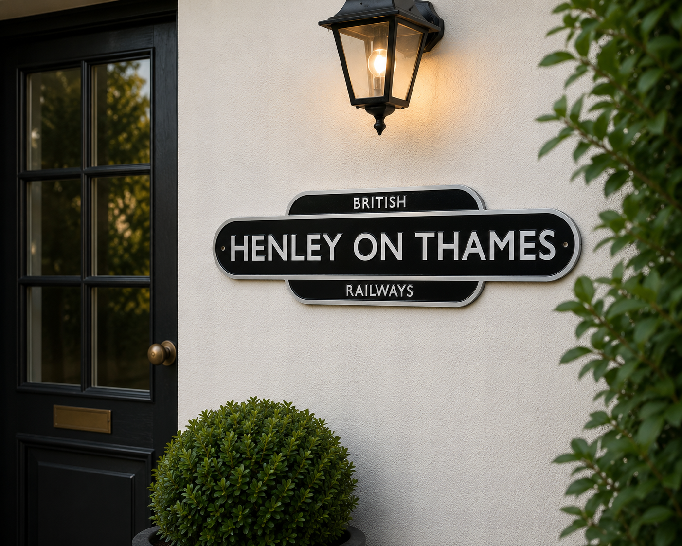 A black and white British Railways station sign reading 'HENLEY ON THAMES' mounted on a cream-colored stucco wall next to a black door and an illuminated wall lamp.