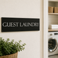 A black rectangular sign with white text reading 'GUEST LAUNDRY' mounted on a beige wall, with an open doorway revealing a laundry room with washing machines, towels, baskets, and plants.