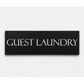 A rectangular black sign with the words 'GUEST LAUNDRY' in white serif lettering, mounted on a textured white wall.