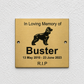 A square brushed gold metal memorial plaque mounted on a textured white wall. The plaque features a black engraved silhouette of a dog and the text 'In Loving Memory of Buster 13 May 2010 - 23 June 2023 R.