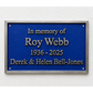 A rectangular blue memorial plaque with a silver border, mounted on a white textured wall. The plaque features raised silver lettering that reads 'In memory of Roy Webb 1936 - 2025 Derek & Helen Bell-Jones'.