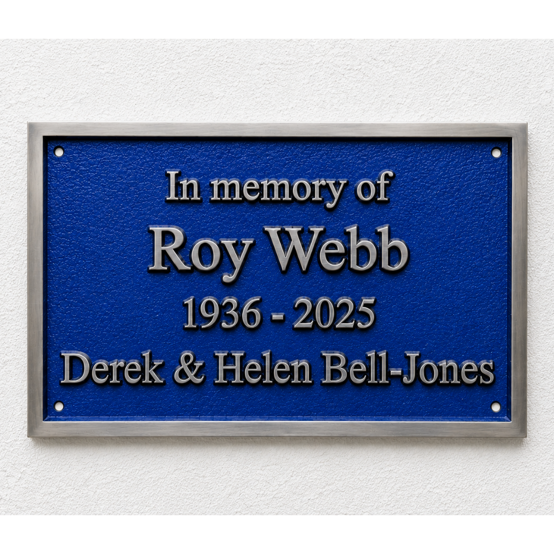 A rectangular blue memorial plaque with a silver border, mounted on a white textured wall. The plaque features raised silver lettering that reads 'In memory of Roy Webb 1936 - 2025 Derek & Helen Bell-Jones'.