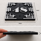 Black metal house number plaque showing dimensions of 14cm by 14cm and 8mm thickness, with the number 48 in silver. The top half shows the plaque mounted on a textured wall, and the bottom half shows a hand holding the plaque to demonstrate its thickness.