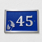 A rectangular blue house number plaque with raised silver numerals '45' and two silver fern motifs, mounted on a white textured wall.