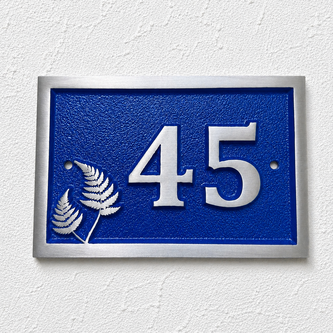 A rectangular blue house number plaque with raised silver numerals '45' and two silver fern motifs, mounted on a white textured wall.