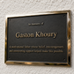 A rectangular black memorial plaque with a polished gold-toned metal border, mounted on a textured off-white wall. The plaque features raised gold lettering that reads 'In memory of Gaston Khoury' and a shorter descriptive quote below.